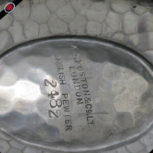 May include: Close-up of a pewter dish with a textured surface. The dish has an oval shape with the text "A.E. POSTON & CO. LTD. LONDON ENGLISH PEWTER 2182" engraved on the surface.