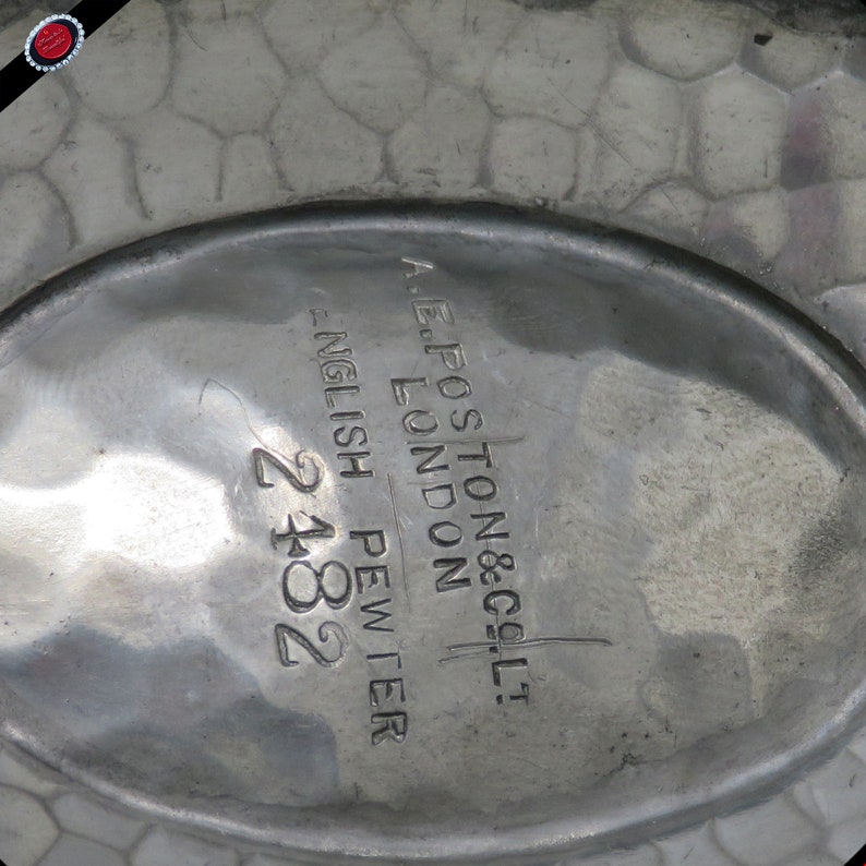 May include: Close-up of a pewter dish with a textured surface. The dish has an oval shape with the text "A.E. POSTON & CO. LTD. LONDON ENGLISH PEWTER 2182" engraved on the surface.
