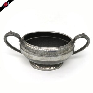 May include: A vintage silver-toned sugar bowl with two handles. The bowl has a hammered texture and a circular base. The handles are curved and attached to the sides of the bowl. The bowl is designed for serving sugar or other condiments.