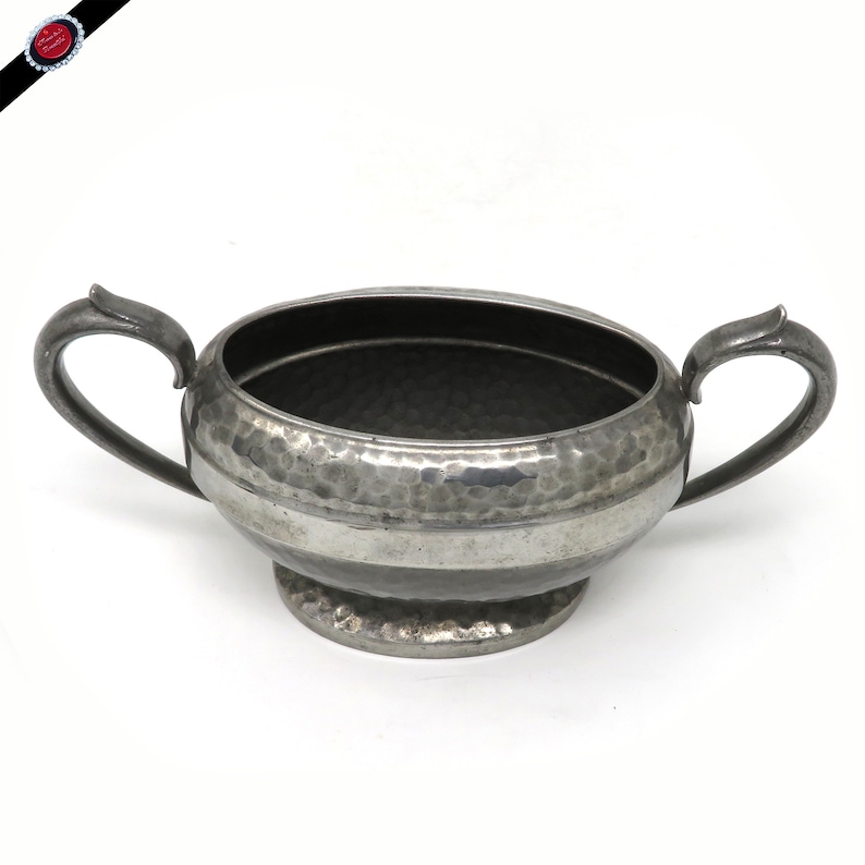 May include: A vintage silver-toned sugar bowl with two handles. The bowl has a hammered texture and a circular base. The handles are curved and attached to the sides of the bowl. The bowl is designed for serving sugar or other condiments.