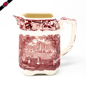May include: A red and white ceramic creamer with a square body and a handle. The creamer features a detailed scene of a building with trees and people in red.