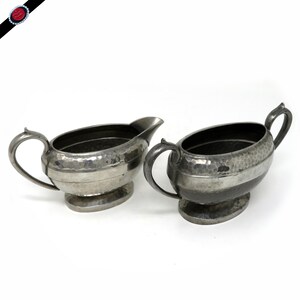 May include: Two antique silver-colored cream and sugar serving dishes. Each piece has a hammered texture and a handle. The cream pitcher has a spout. Both sit on oval bases. The set is likely vintage.