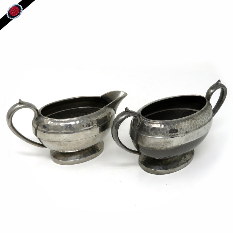 May include: Two antique silver-colored cream and sugar serving dishes. Each piece has a hammered texture and a handle. The cream pitcher has a spout. Both sit on oval bases. The set is likely vintage.