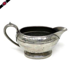 May include: A silver-colored creamer with a hammered texture. The creamer has a curved handle, a spout, and a round base. The interior is dark. The creamer is likely made of metal and is designed for serving liquids.