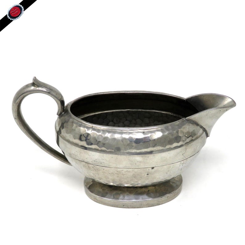 May include: A silver-colored creamer with a hammered texture. The creamer has a curved handle, a spout, and a round base. The interior is dark. The creamer is likely made of metal and is designed for serving liquids.