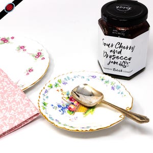 May include: A white porcelain plate with a floral design and a gold rim. A silver spoon is resting on the plate. A jar of Sour Cherry and Prosecco Jam is in the background. The jar has a white label with black text that reads "Sour Cherry and Prosecco Jam" and "Wickstead Relish Company".