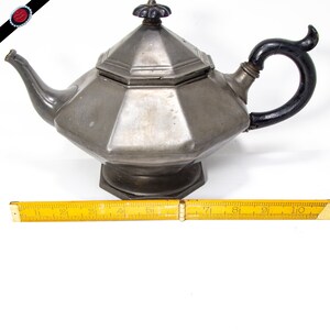 May include: A silver teapot with a black handle and a octagonal body. The teapot has a lid with a decorative knob. The teapot is sitting on a white surface.
