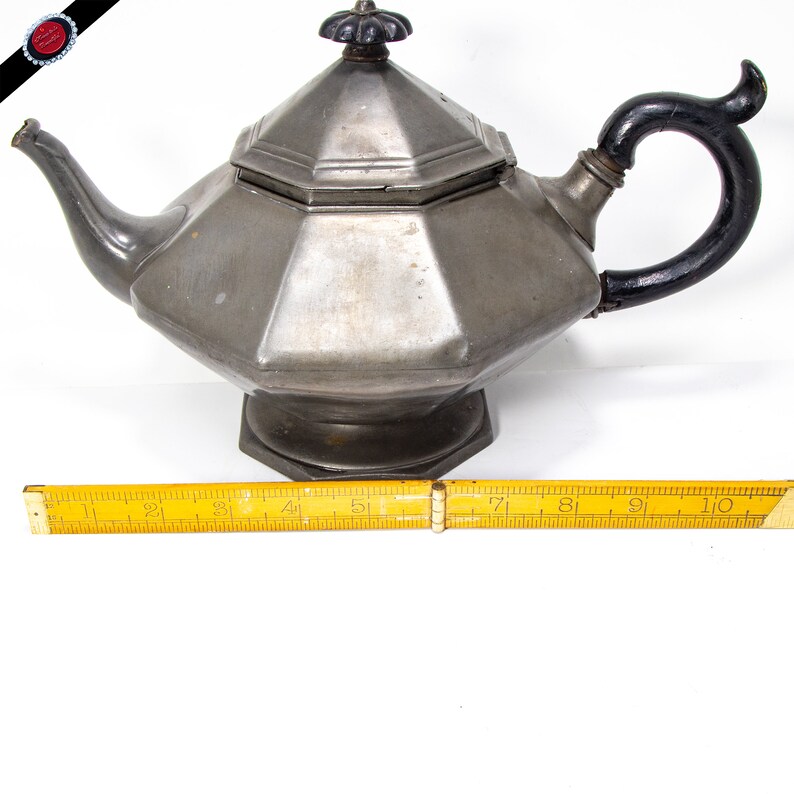 May include: A silver teapot with a black handle and a octagonal body. The teapot has a lid with a decorative knob. The teapot is sitting on a white surface.