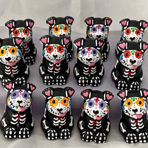 Puede incluir: Doce figuritas de perro de cer&aacute;mica negra y blanca con dise&ntilde;os coloridos de calaveras de az&uacute;car. Cada figurita tiene un dise&ntilde;o diferente y presenta un coraz&oacute;n rosa en cada oreja.