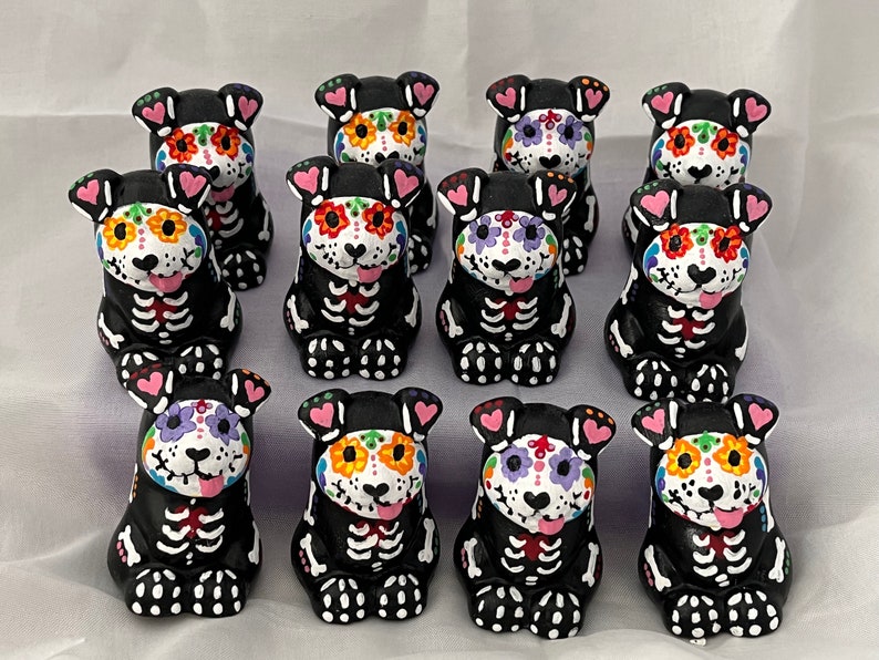 Puede incluir: Doce figuritas de perro de cer&aacute;mica negra y blanca con dise&ntilde;os coloridos de calaveras de az&uacute;car. Cada figurita tiene un dise&ntilde;o diferente y presenta un coraz&oacute;n rosa en cada oreja.
