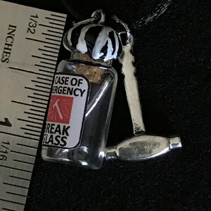 May include: A novelty pendant featuring a miniature glass bottle with a cork stopper, labeled "CASE OF EMERGENCY" and "BREAK GLASS." The bottle is paired with a small silver hammer. The bottle's cap is painted with black and white stripes.