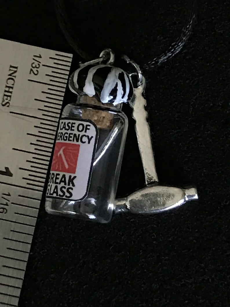 May include: A novelty pendant featuring a miniature glass bottle with a cork stopper, labeled "CASE OF EMERGENCY" and "BREAK GLASS." The bottle is paired with a small silver hammer. The bottle's cap is painted with black and white stripes.