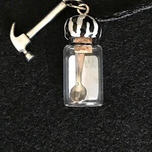 May include: A clear glass vial pendant with a silver spoon inside, topped with a black and white striped cap and a silver hammer charm. The pendant hangs from a black cord.