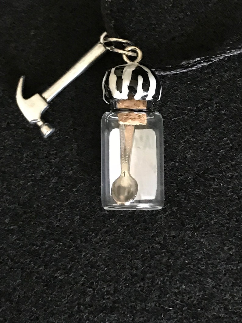 May include: A clear glass vial pendant with a silver spoon inside, topped with a black and white striped cap and a silver hammer charm. The pendant hangs from a black cord.