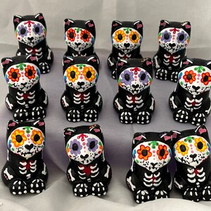 Puede incluir: Una colecci&oacute;n de 10 figuritas de gatos negros con dise&ntilde;os coloridos de calaveras de az&uacute;car. Cada gato est&aacute; pintado con un esquema de color diferente, incluyendo rosa, morado, naranja y amarillo. Los gatos est&aacute;n sentados en fila sobre una superficie blanca.