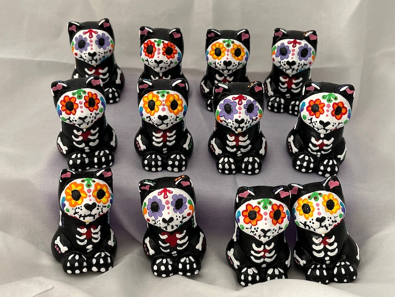 Puede incluir: Una colecci&oacute;n de 10 figuritas de gatos negros con dise&ntilde;os coloridos de calaveras de az&uacute;car. Cada gato est&aacute; pintado con un esquema de color diferente, incluyendo rosa, morado, naranja y amarillo. Los gatos est&aacute;n sentados en fila sobre una superficie blanca.