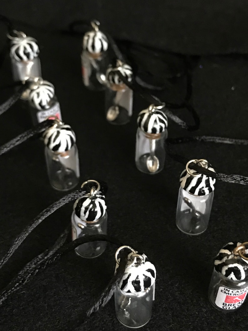 May include: A collection of small glass vial necklaces with black cords. Each vial has a decorative lid with a black and white zebra stripe pattern. Some vials have small silver spoons inside. The background is black.