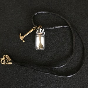 May include: A black cord necklace featuring a small glass vial pendant with a spoon inside. A miniature gold-colored hammer charm and a decorative clasp complete the necklace. The background is a dark, textured surface.
