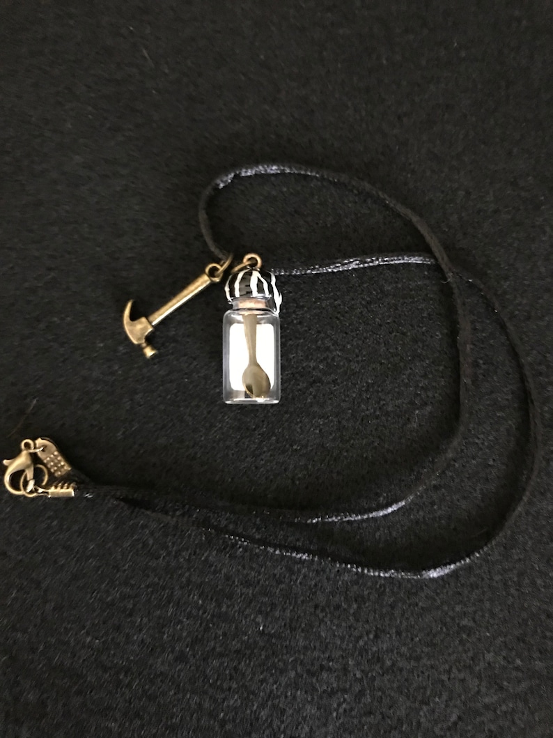 May include: A black cord necklace featuring a small glass vial pendant with a spoon inside. A miniature gold-colored hammer charm and a decorative clasp complete the necklace. The background is a dark, textured surface.