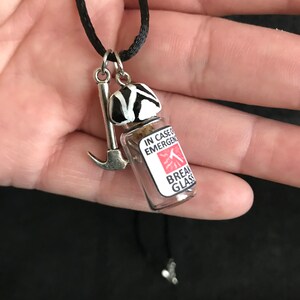 May include: A novelty necklace with a black cord. The pendant features a small glass vial with the text "IN CASE OF EMERGENCY BREAK GLASS", a miniature ice axe, and a black and white striped object. The necklace is held in a hand.
