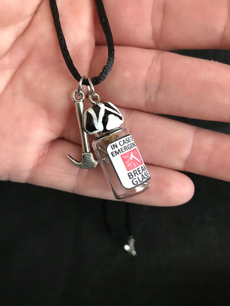 May include: A novelty necklace with a black cord. The pendant features a small glass vial with the text "IN CASE OF EMERGENCY BREAK GLASS", a miniature ice axe, and a black and white striped object. The necklace is held in a hand.