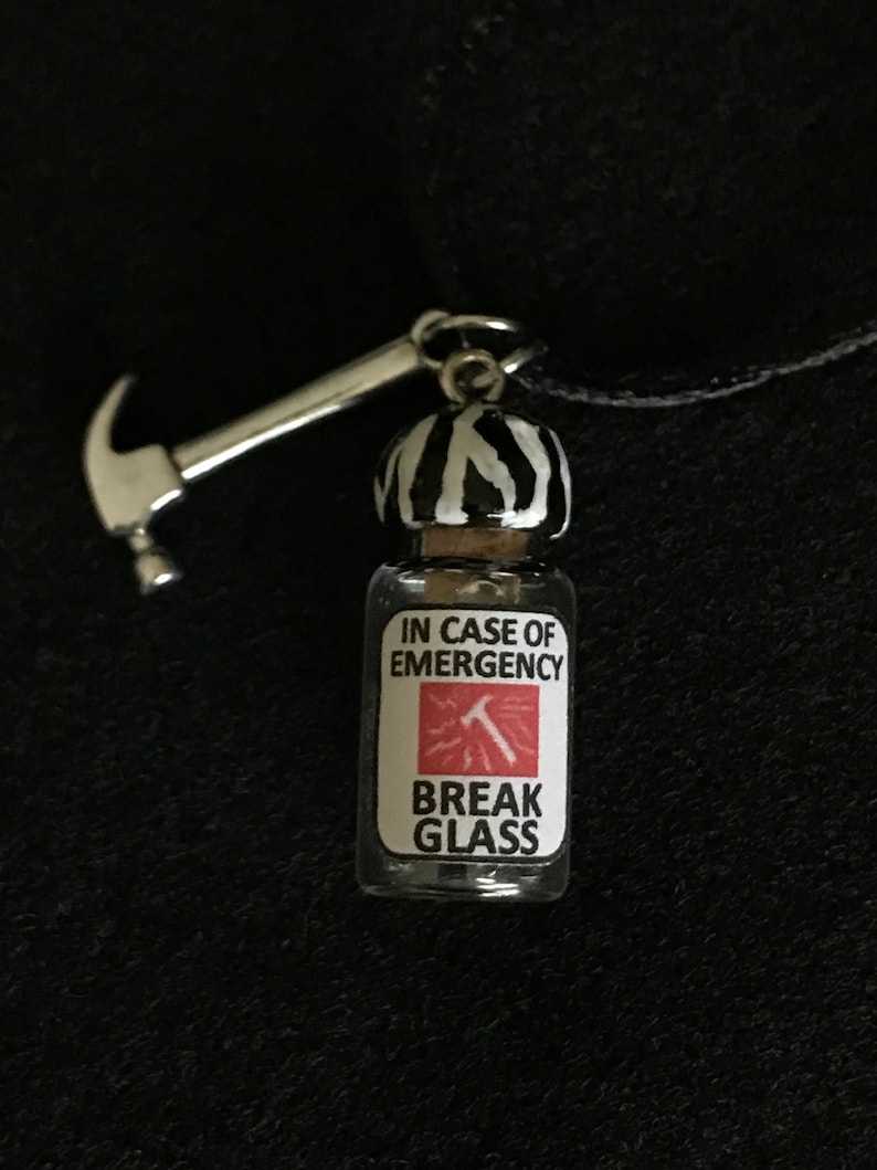 May include: A novelty pendant necklace featuring a small glass bottle with a black and white striped top and a silver hammer charm. The bottle has a label that reads "IN CASE OF EMERGENCY BREAK GLASS."
