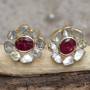 以下が含まれることがあります： ゴールドトーンのスタッドピアス。各イヤリングは、中央に楕円形の濃い赤色の宝石があり、その周りを不規則な形の透明な宝石のハローで囲んでいます。 イヤリングは木製の表面に表示されています。