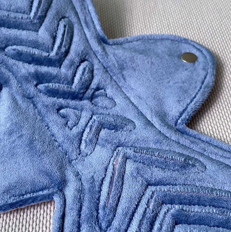 Op de afbeelding: Close-up van een lichtblauw, herbruikbaar maandverband. Het maandverband heeft een ingewikkeld geborduurd ontwerp en een drukknoopsluiting. De zachte, absorberende stof is zichtbaar en toont de textuur en constructie van het maandverband. Het maandverband is ontworpen voor comfort en milieuvriendelijkheid.