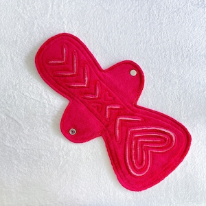 Puede incluir: Una compresa menstrual reutilizable de color rosa brillante con costuras decorativas. La compresa presenta un diseño de corazón y flecha, con broches para un cierre seguro. La compresa está hecha de un material suave y absorbente.