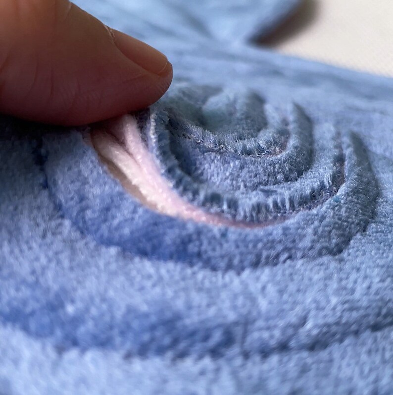 Op de afbeelding: Close-up van een lichtblauwe, zachte stof met een spiraalvormig ontwerp. Het ontwerp is geborduurd met een lichtere blauwe draad en een roze accent is zichtbaar in de spiraal. De stof heeft een zachte, getextureerde uitstraling.