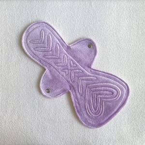 Puede incluir: Una compresa menstrual reutilizable de color lavanda con un diseño de corazón y flecha. La compresa tiene una superficie suave y texturizada y se asegura con dos broches. El diseño está bordado con un tono más claro de lavanda.