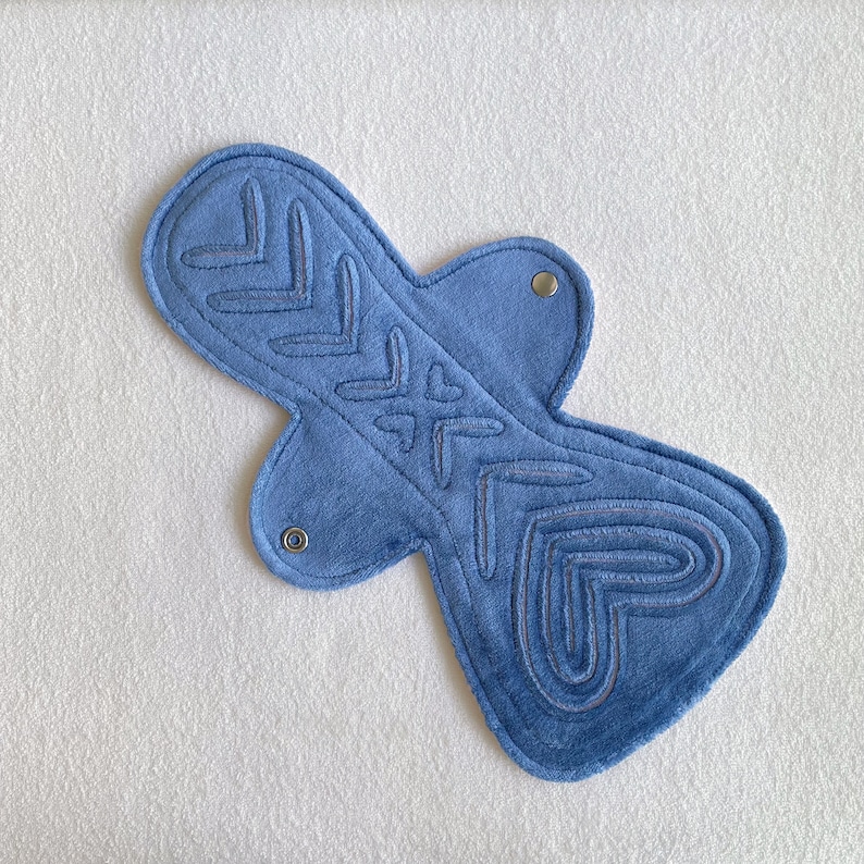 Op de afbeelding: Een blauw herbruikbaar maandverband met een decoratief gestikt ontwerp. Het maandverband heeft gestikte pijlen en hartvormen. Metalen drukknopen zijn zichtbaar op de vleugels.