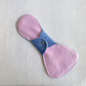Op de afbeelding: Een herbruikbaar stoffen maandverband met een lichtroze absorberend oppervlak en een blauwe achterkant. Het maandverband wordt vastgezet met een drukknoopsluiting. Het is ontworpen voor vrouwelijke hygi&euml;ne en is gemaakt van zachte, absorberende materialen.