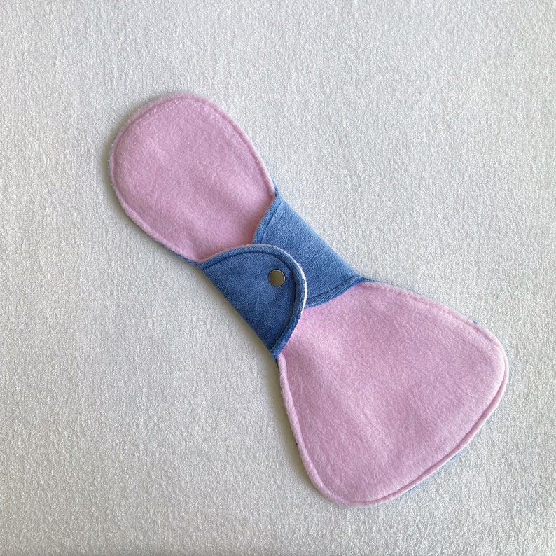 Op de afbeelding: Een herbruikbaar stoffen maandverband met een lichtroze absorberend oppervlak en een blauwe achterkant. Het maandverband wordt vastgezet met een drukknoopsluiting. Het is ontworpen voor vrouwelijke hygi&euml;ne en is gemaakt van zachte, absorberende materialen.