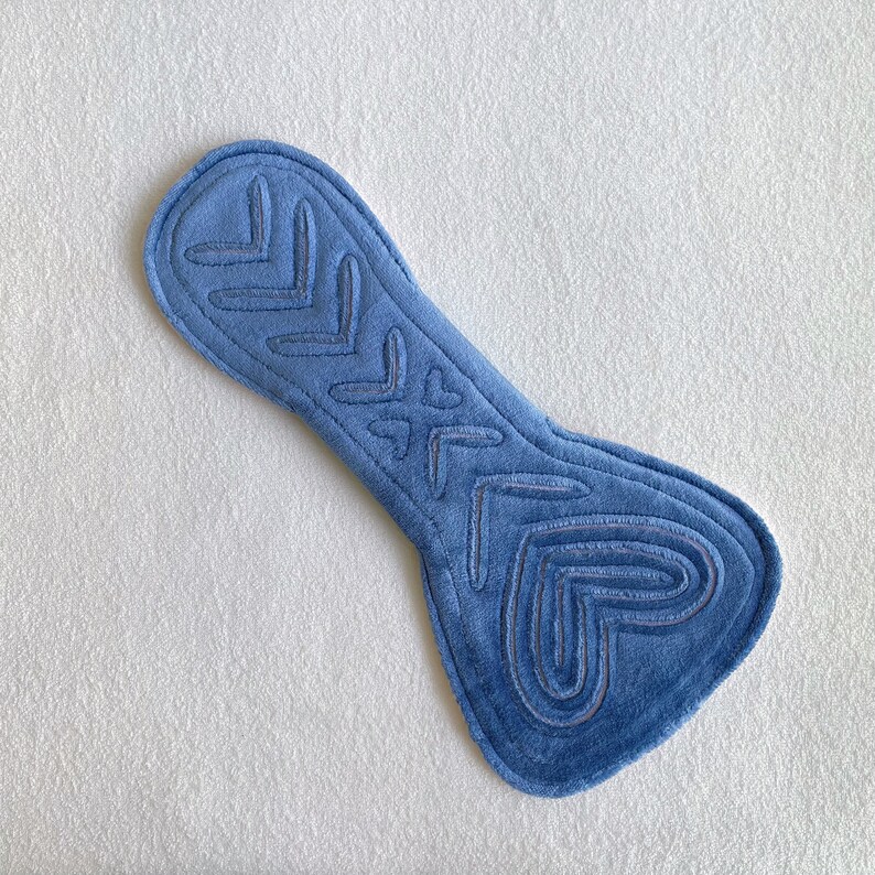Op de afbeelding: Een blauw, op su&egrave;de lijkend kussen met geborduurde hart- en pijlenpatronen. Het kussen heeft een gebogen vorm, breder aan de basis en smaller aan de bovenkant. De borduursels zijn in een donkerdere blauwtint.