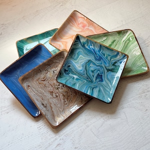May include: Five square trays with a marbled design. The trays are made of wood and have a gold trim. The trays are stacked on top of each other. The colors of the trays are blue, green, brown, and peach.