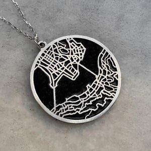 Op de afbeelding: Zilverkleurige ketting met een ronde hanger met een gedetailleerd kaartontwerp. De kaart is zwart, met zilveren straten en contouren. De ketting is delicaat en zilverkleurig, wat een eenvoudig maar elegant accessoire creëert. De hanger is ongeveer 3 cm in diameter.
