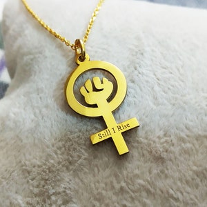 Op de afbeelding: Goudkleurige hangerketting met een symbool van vrouwelijke empowerment. De hanger heeft een vuist in een cirkel, met een kruis eronder. De woorden "Still I Rise" zijn gegraveerd op het kruis. De ketting wordt getoond op een witte, zachte ondergrond.