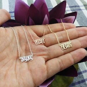 May include: Three gold necklaces with Chinese characters hanging from them. The necklaces are on a hand with a purple background.