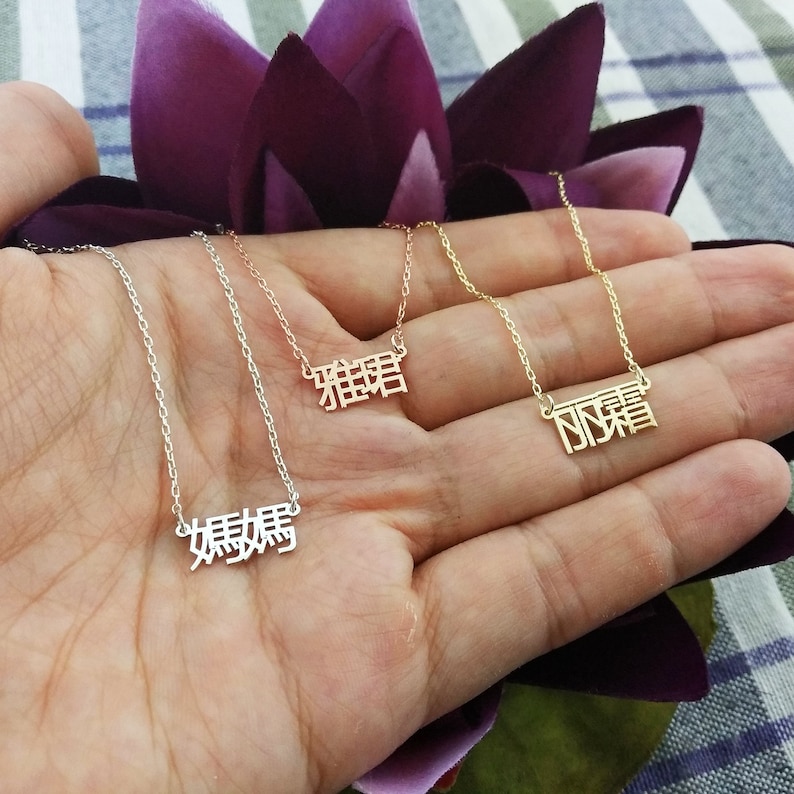 May include: Three gold necklaces with Chinese characters hanging from them. The necklaces are on a hand with a purple background.