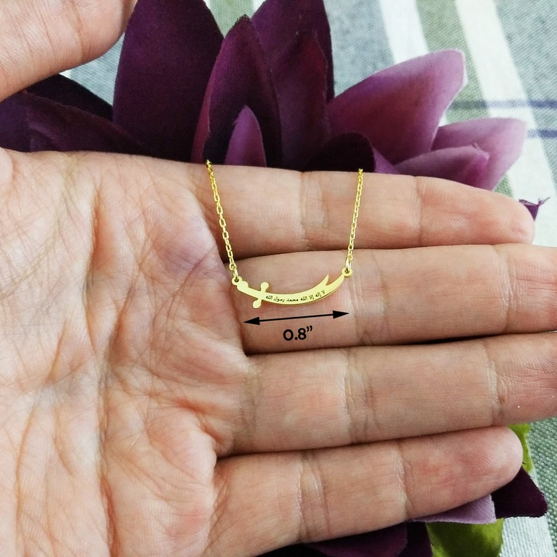May include: Gold necklace with a curved pendant featuring Arabic calligraphy. The pendant measures 0.8 inches in length.