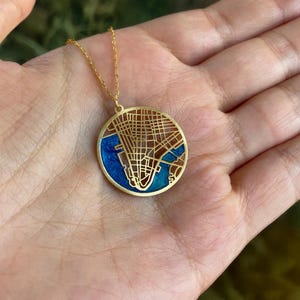 May include: Gold-colored pendant necklace featuring a detailed map design. The circular pendant has a blue enamel background with a gold-colored overlay depicting a city map. The necklace is displayed on a hand.
