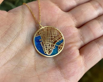 May include: Gold-colored pendant necklace featuring a detailed map design. The circular pendant has a blue enamel background with a gold-colored overlay depicting a city map. The necklace is displayed on a hand.