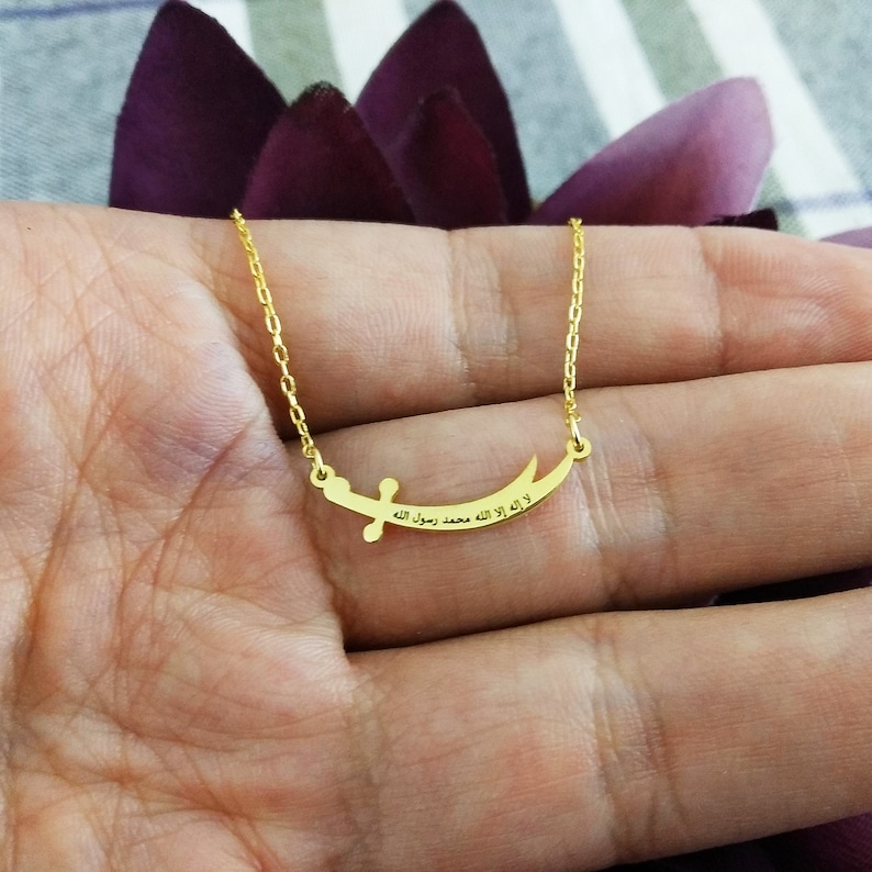 May include: A gold necklace with a crescent moon pendant featuring an inscription in Arabic script. The inscription reads "لا إله إلا الله محمد رسول الله".
