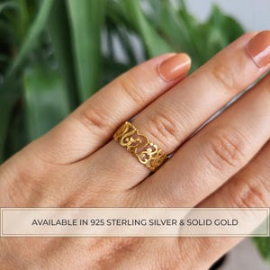 Op de afbeelding: Een goudkleurige ring met een ingewikkeld, opengewerkt ontwerp met herhalende cursieve letters. De band heeft een gepolijste afwerking en een doorlopend patroon. De tekst onderaan luidt: AVAILABLE IN 925 STERLING SILVER & SOLID GOLD.
