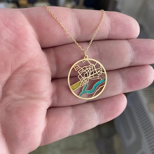 May include: A gold-colored necklace featuring a circular pendant with a detailed map design. The map includes streets and waterways, with sections colored in teal and yellow. The necklace is held in a hand.