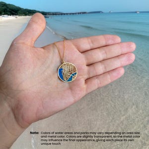 May include: A gold-colored pendant necklace featuring a detailed map design of a city, with blue and brown accents. The pendant is round and held in a hand, with a beach and ocean in the background.
