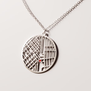 May include: Silver-colored pendant necklace featuring a circular map design. The map has a grid pattern with a small red dot. The pendant hangs from a delicate chain, creating a minimalist and modern aesthetic.