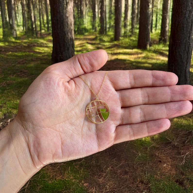 May include: A gold-colored pendant necklace with a circular design. The pendant features a geometric pattern with clear and green sections. The necklace is held in an open hand against a backdrop of a forest.