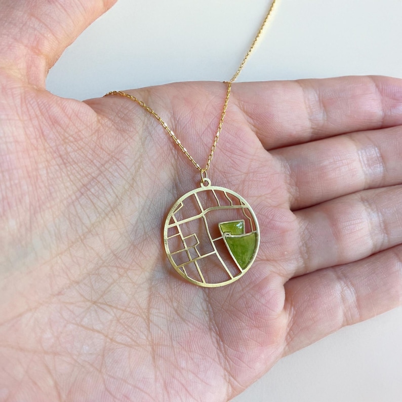 May include: A gold-colored necklace with a circular pendant featuring a geometric design and a green accent. The pendant hangs from a delicate gold chain. The necklace is displayed on a hand.